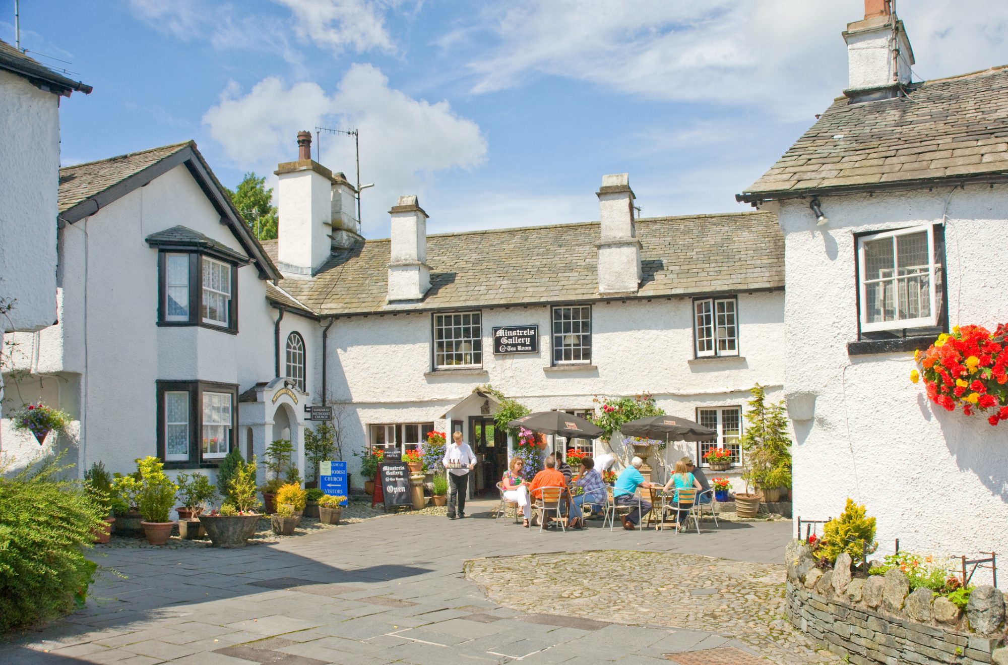 An image of the Minstrels Gallery Tea Room at Hawkshead in the English Lake District with customers dining and drinking outside. The image aims to convey a holiday atmosphere in this attractive tourist destination.